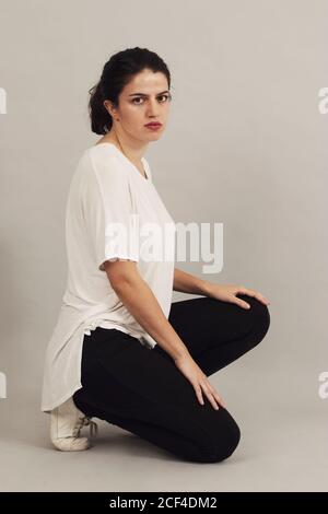 Side view of pensive youthful dark haired female in sportswear looking at camera while crouching on gray background in studio Stock Photo