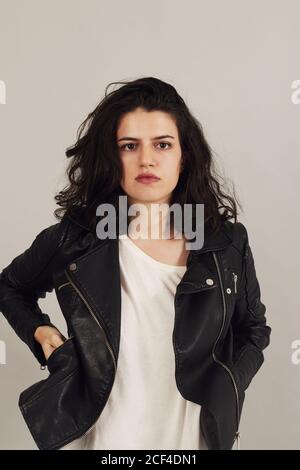 Thoughtful attractive dark haired Woman in black biker jacket and looking at camera on gray background in studio Stock Photo