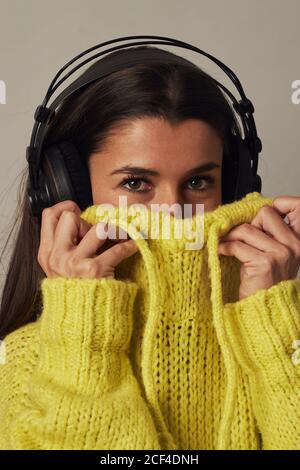 Cheerful young dark haired female in headphones looking at camera while covering face with yellow cozy sweater on gray background in studio Stock Photo