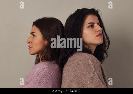 Side view of satisfied young charming dark haired women wearing sweaters and looking at camera while standing together on gray background in studio Stock Photo