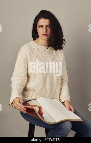 Serious youthful dark haired Woman in glasses and white sweater looking at camera holding a magazine while sitting in studio Stock Photo