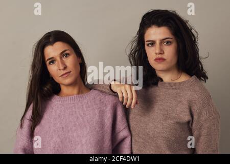 Satisfied young charming dark haired women wearing sweaters and looking at camera while standing together on gray background in studio Stock Photo