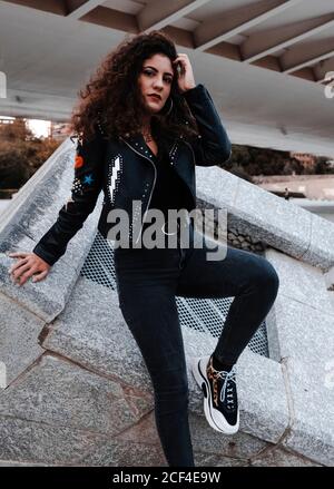 From below of stylish confident long haired brunette wearing rocker styled black leather jacket with stripes and skinny jeans with trendy sneakers standing next to concrete construction in city and looking at camera Stock Photo