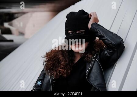 Young Woman in black mask wearing black leather jacket with colorful and metal stripes standing on street leaning back on concrete wall and looking at camera Stock Photo