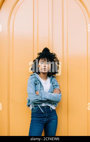 Young African American Woman in jeans and denim jacket leaning on yellow door and looking at camera Stock Photo