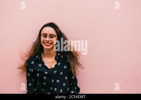 Charming teen Woman in eyeglasses and blue shirt smiling happily at camera standing against pink wall Stock Photo