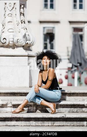 Beautiful ethnic Woman in jeans and tank top relaxing and sunbathing on stone stairs against urban background Stock Photo