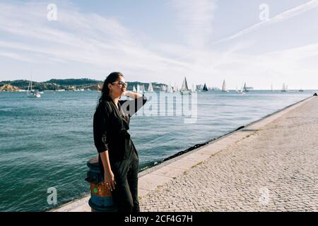Side view of young attractive Woman in black outfit standing by sea and leaning on pole in Lisbon Stock Photo
