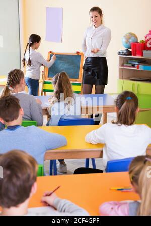 Positive friendly  diligent  female teacher standing at blackboard with schoolgirl during lesson in classroom Stock Photo
