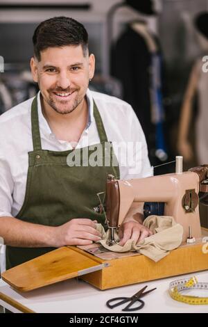 Diligent focused male tailor in apron sewing outfit details using modern sewing machine at table while creating exclusive clothes collection in contemporary work studio Stock Photo
