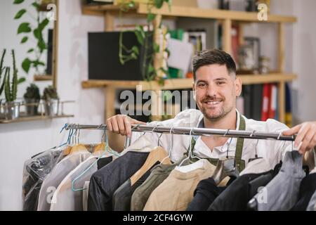 High angle of happy focused male tailor in apron checking details of apparel hanging on hanger on metal rack among other trendy bespoke clothes in modern workroom Stock Photo