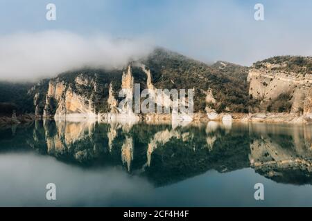 Magnificent landscape of calm lake with mirrored water surface surrounded by rough rocky mountains of Montsec Range covered with dense fog in cold day in Spain Stock Photo