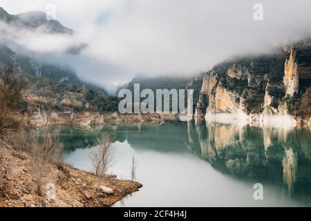 Magnificent landscape of calm lake with mirrored water surface surrounded by rough rocky mountains of Montsec Range covered with dense fog in cold day in Spain Stock Photo