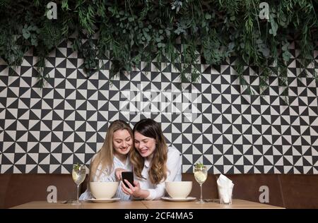 Elegant women friends using phone while sitting at table of restaurant during lunch Stock Photo
