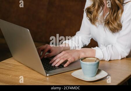 Crop view of anonymous female freelancer browsing modern laptop while sitting at table with a cup of coffee in cozy restaurant Stock Photo