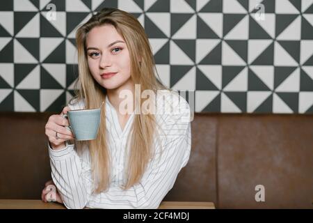 Pensive Woman with cup of fresh coffee looking at camera while sitting at table in cozy restaurant Stock Photo