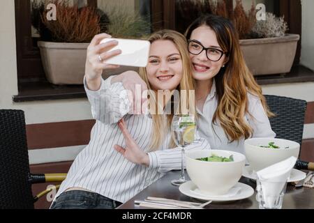 Two stylish female friends smiling and posing for selfie while sitting at table in cozy restaurant Stock Photo