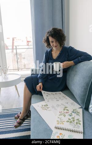 Beautiful young brunette female sitting on sofa and looking album with drawings at home while enjoying a cup of coffee Stock Photo
