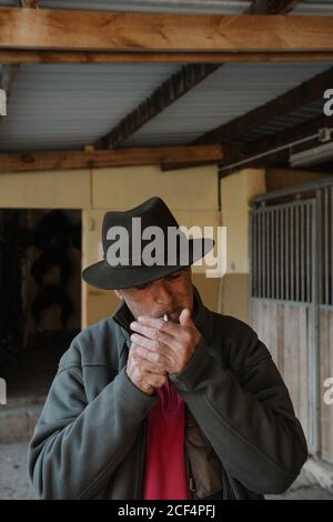 Adult male in hat smoking cigarette while standing near stalls inside stable on ranch Stock Photo