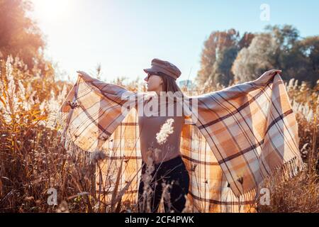 Autumn fashion. Young woman wearing stylish poncho, cap outdoors raising hands. Clothing and accessories. Stock Photo