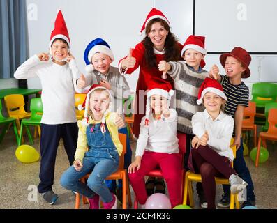 Cheerful group of pupils with female teacher wearing Santa hats posing together in schoolroom, giving thumbs up Stock Photo