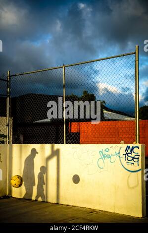 Shadows on concrete wall of anonymous man and kid playing ball on playground in sun rays under blue sky with gray lush dramatic clouds Stock Photo