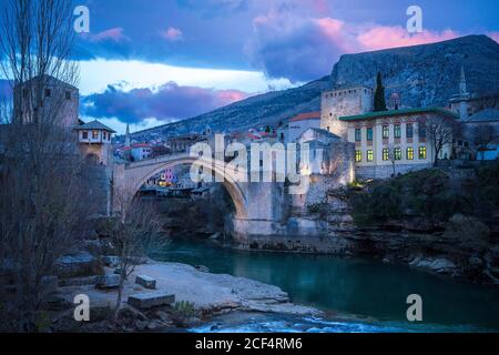 Wonderful scenery of tranquil turquoise river against historical district of picturesque city Mostar with medieval bridge and old buildings at foot of mountain under colorful cloudy sky during sunrise in Federation of Bosnia and Herzegovina Stock Photo