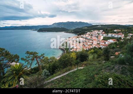 Drone view of peaceful seashore with green trees and small town with red roofs locating near blue sea and mountain ridge against blue cloudy sky Stock Photo