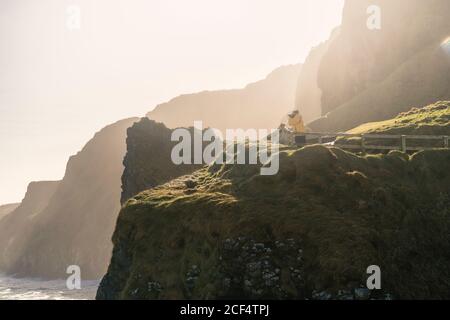 Side view of man traveler in warm activewear standing on rural road running along shore with green grass and taking photo with professional camera of ocean with calm water in sunny spring day in Northern Ireland Stock Photo