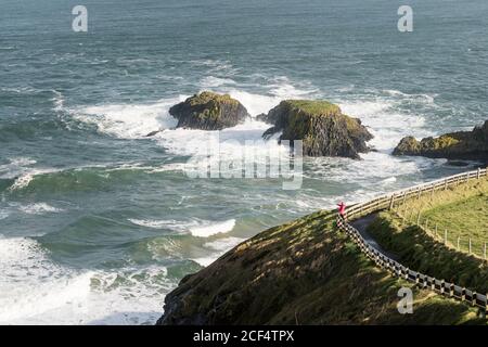 Aerial view of unrecognizable person walking on narrow rural road running along green field on sunny spring day at coastline in Northern Ireland Stock Photo