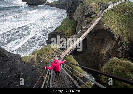 Back view of unrecognizable Woman tourist in red jacket walking on rope Carric a rede bridge suspended between mainland and small rocky island at Northern Ireland coastline