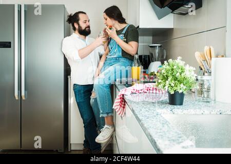 Bearded man feeding girlfriend with healthy food while spending time on kitchen at home together Stock Photo