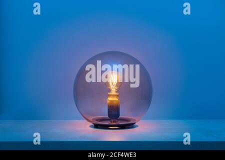 Modern turned on luminaire with light bulb inside thin transparent glass sphere in middle of shelf in blue room at dusk Stock Photo