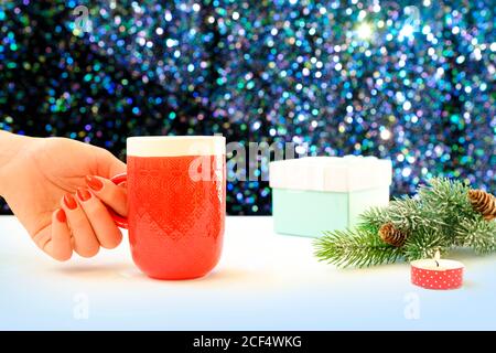 Woman's hand holding a mug of coffee on a Christmas background. Christmas gift boxes and green fir tree over shining table. Stock Photo