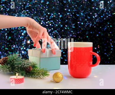 Woman's hand holding a gift box on a Christmas background. Red cup of coffee and green spruce over a shiny table. Stock Photo