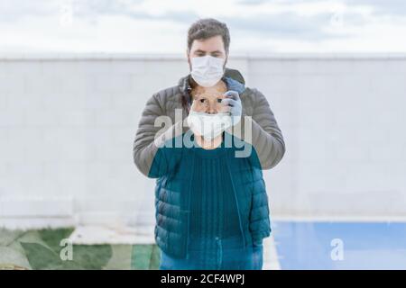 Dark haired young male in warm jacket and medical mask taking photo of senior Woman in medical mask through window while standing outside in front of modern hospital Stock Photo