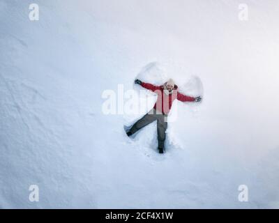 From above smiling middle-aged male in outwear lying on white snow and making snow angel silhouette Stock Photo