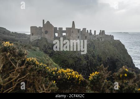 Majestic landscape with ruins of ancient stone Dunluce Castle in sunlight located on edge of cliff with rainbow breaking through stormy sky and sea waves beating against shore Stock Photo