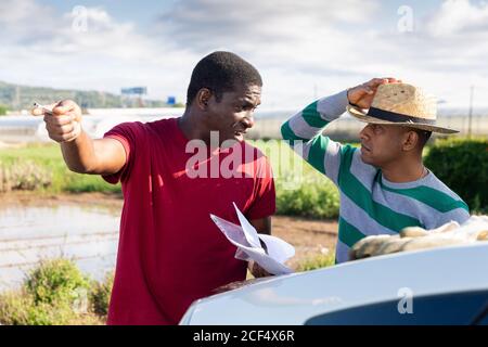 Disgruntled african american farmer holding papers, reprimanding hispanic contractor for poor job outdoors Stock Photo