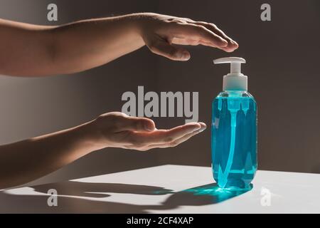Crop faceless person pulling hands for plastic blue liquid soap bottle with white dispenser inside located on white table with shadow Stock Photo