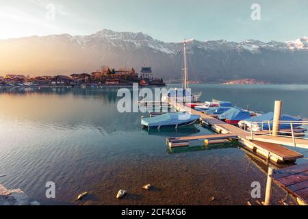 Landscape of peaceful blue lake with small pier and boats on background of mountains in sunshine, Switzerland Stock Photo