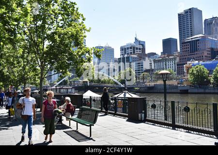 People walk along the Southbank Promenade in downtown Melbourne on a bright sunny day.  The Yarra river can be seen flowing through the city centre. Stock Photo