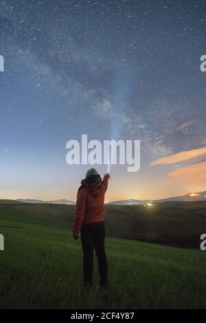 Back view of traveler standing in green highlands and glowing with torch in starry sky, Tuscany, Italy Stock Photo