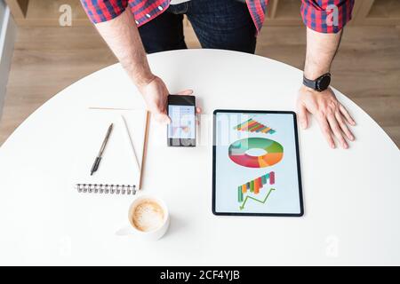 An anonymous man is holding a mobile phone. The man is working from home. The tablet and the phone show graphics. There is a mug of coffee in the desk Stock Photo