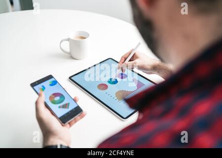 An anonymous man is writing on a tablet with graphics. The man is working from home. The entrepreneur is holding a phone with graphics from above Stock Photo