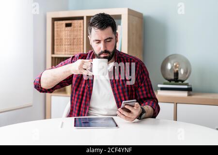 A young man is looking at the mobile phone. The man is working from home. The entrepreneur is holding a phone and drinking from a mug Stock Photo