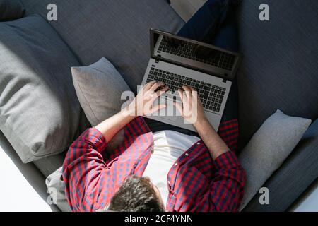 An anonymous man is working from home. The entrepreneur is typing on a computer from above. He is sitting on the sofa Stock Photo