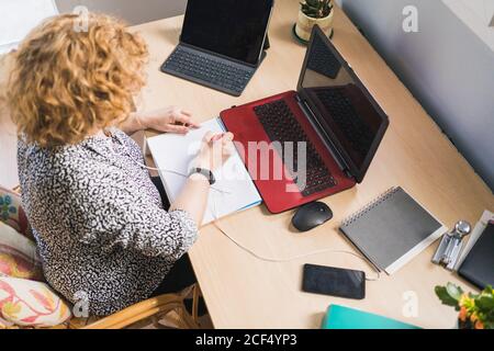 From above view of anonymous female freelancer taking notes with pen in notebook while listening music in earphones on smartphone and sitting at desk with gadgets at home Stock Photo