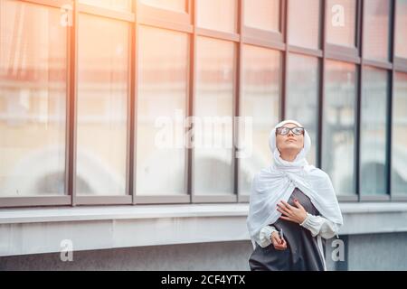 Alone muslim girl in white hijab, with glasses walking and looking in the sky. near building. outdoor Stock Photo