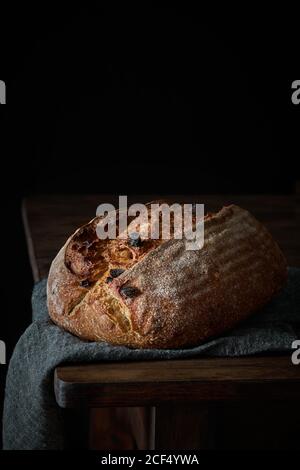 Crispy freshly baked sourdough bread loaf with raisin placed on gray cloth on wooden table with black background Stock Photo